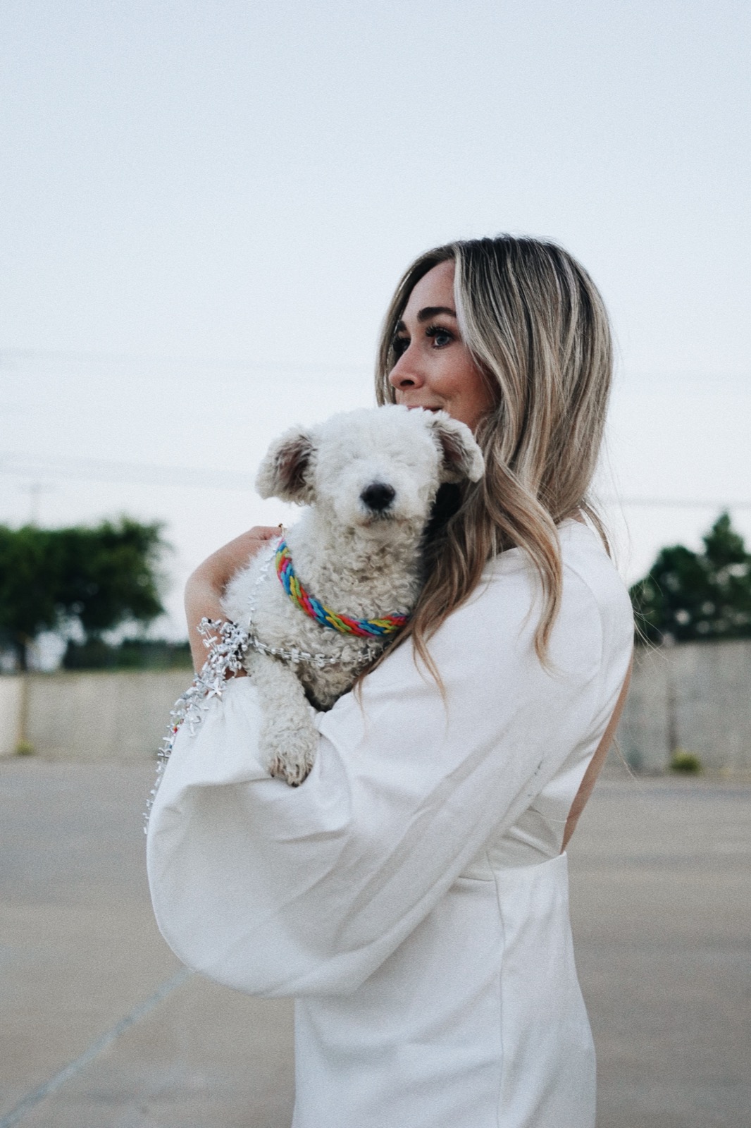 Mia holding Phoebe, a rescued white poodle, in a white dress at dusk